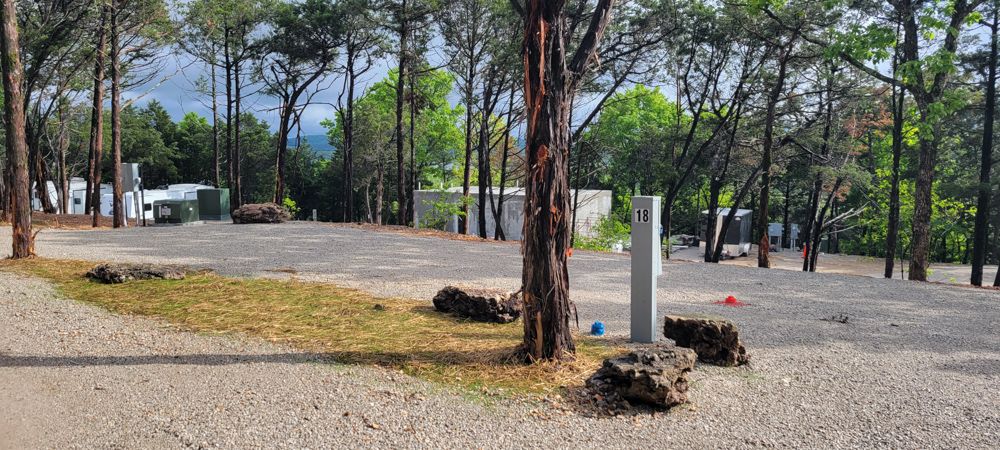 Open gravel RV site number 18 framed by tall pines, with a distant view of the Ozark hills.