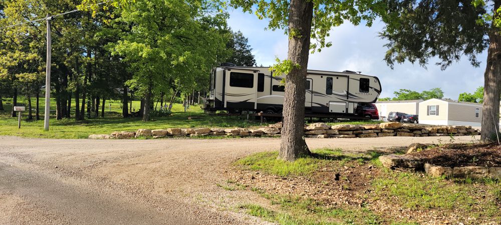 A large fifth-wheel RV parked on a lakeview site edged with a natural stone retaining wall.